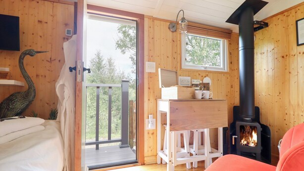 Interior acolhedor de uma cabana, com decoração em madeira, uma cama, uma escrivaninha, uma cadeira e uma lareira a lenha.