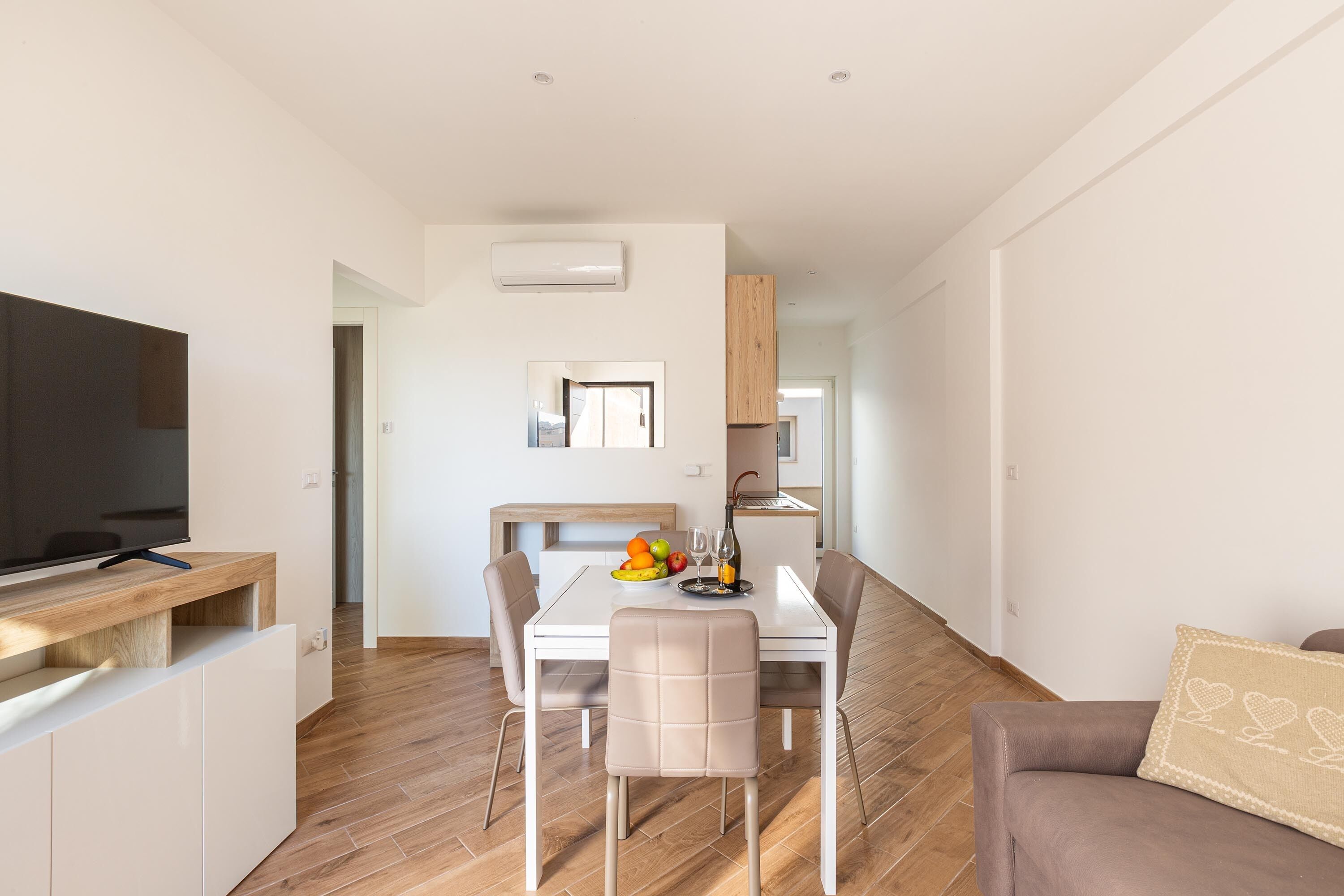 Una sala de estar moderna con un televisor de pantalla plana, una mesa de comedor con sillas, un sofá y una zona de cocina al fondo.