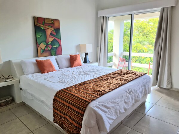 Habitación de hotel con una cama grande, un cuadro en la pared, un balcón con vistas a la vegetación y una silla afuera.