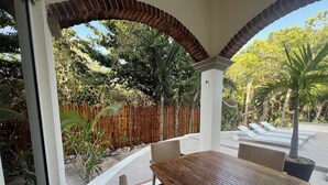 Un patio al aire libre con una mesa y sillas de madera, rodeado por una valla de bambú, palmeras y vistas a un jardín.