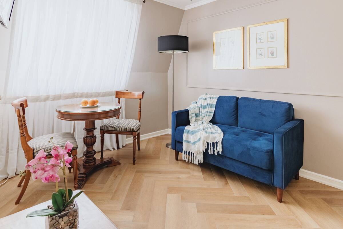 Ein Wohnzimmer mit einem blauen Sofa, einem runden Holztisch mit zwei Stühlen, einer Stehlampe und einer Vase mit rosa Blumen.