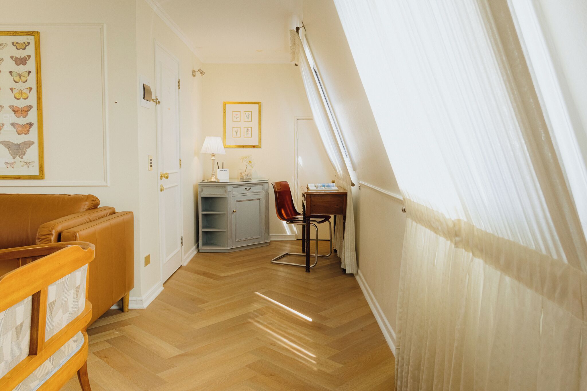 Une pièce avec un plancher en bois, un canapé en cuir marron, un bureau en bois et une chaise. Un tableau encadré représentant un papillon est accroché au mur, et un autre tableau encadré se trouve au-dessus du bureau.