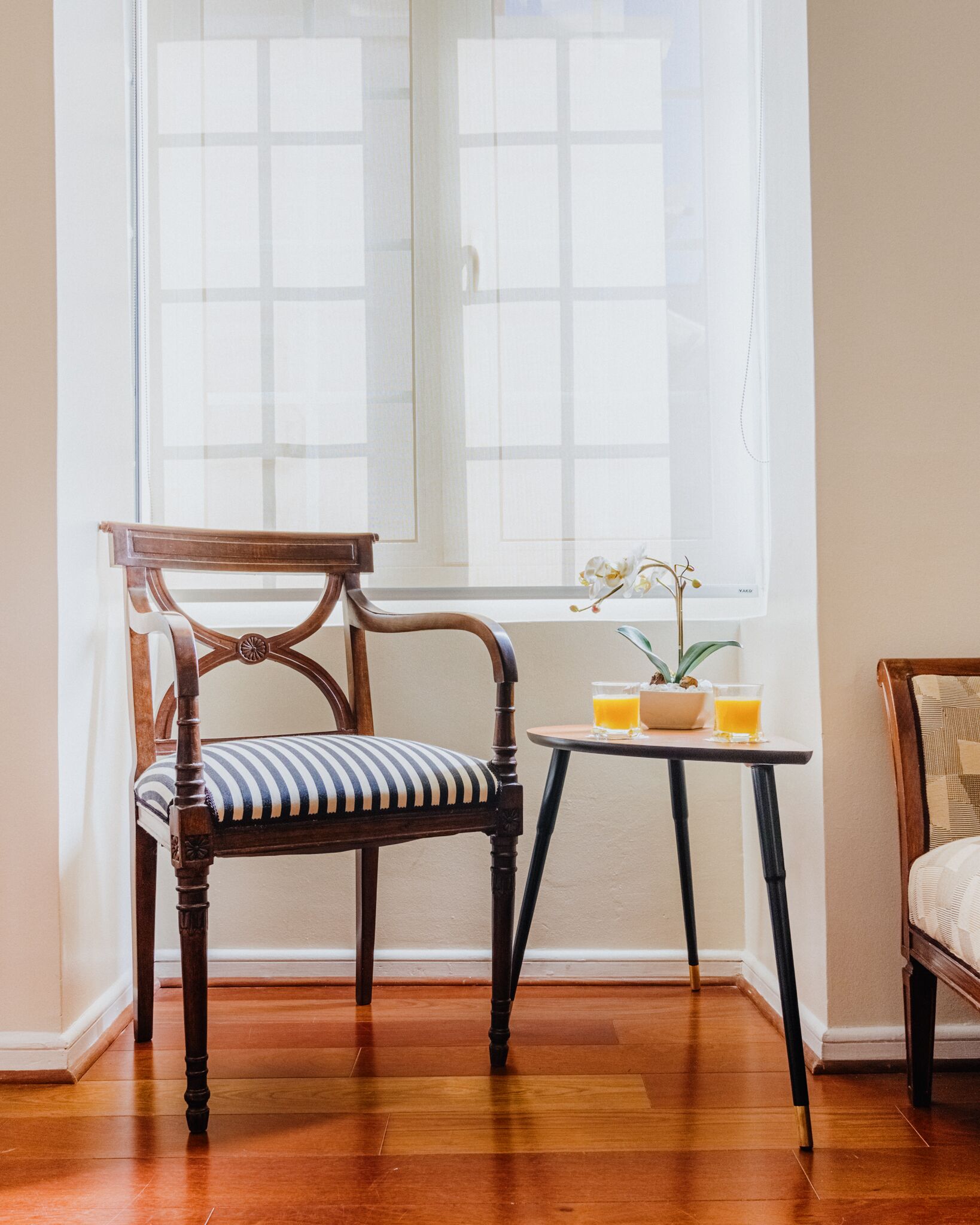 Una silla de madera con cojín a rayas, una mesita con planta en maceta y dos vasos de jugo de naranja, y una ventana con vistas al exterior.