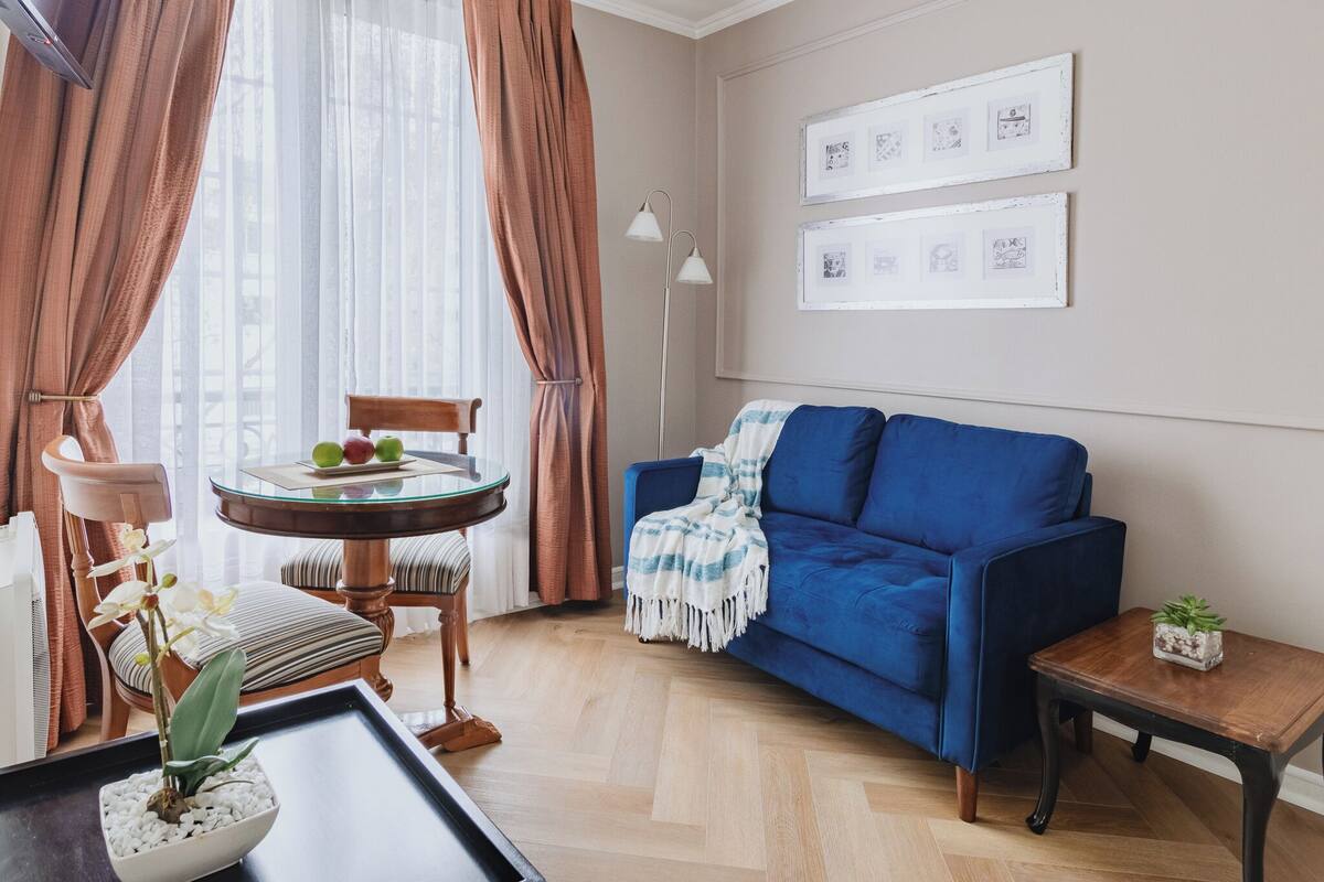 Ein Wohnzimmer mit einem blauen Sofa, einem runden Couchtisch mit Früchten, einem kleinen Tisch mit einer Pflanze und einer Wand mit gerahmten Bildern.