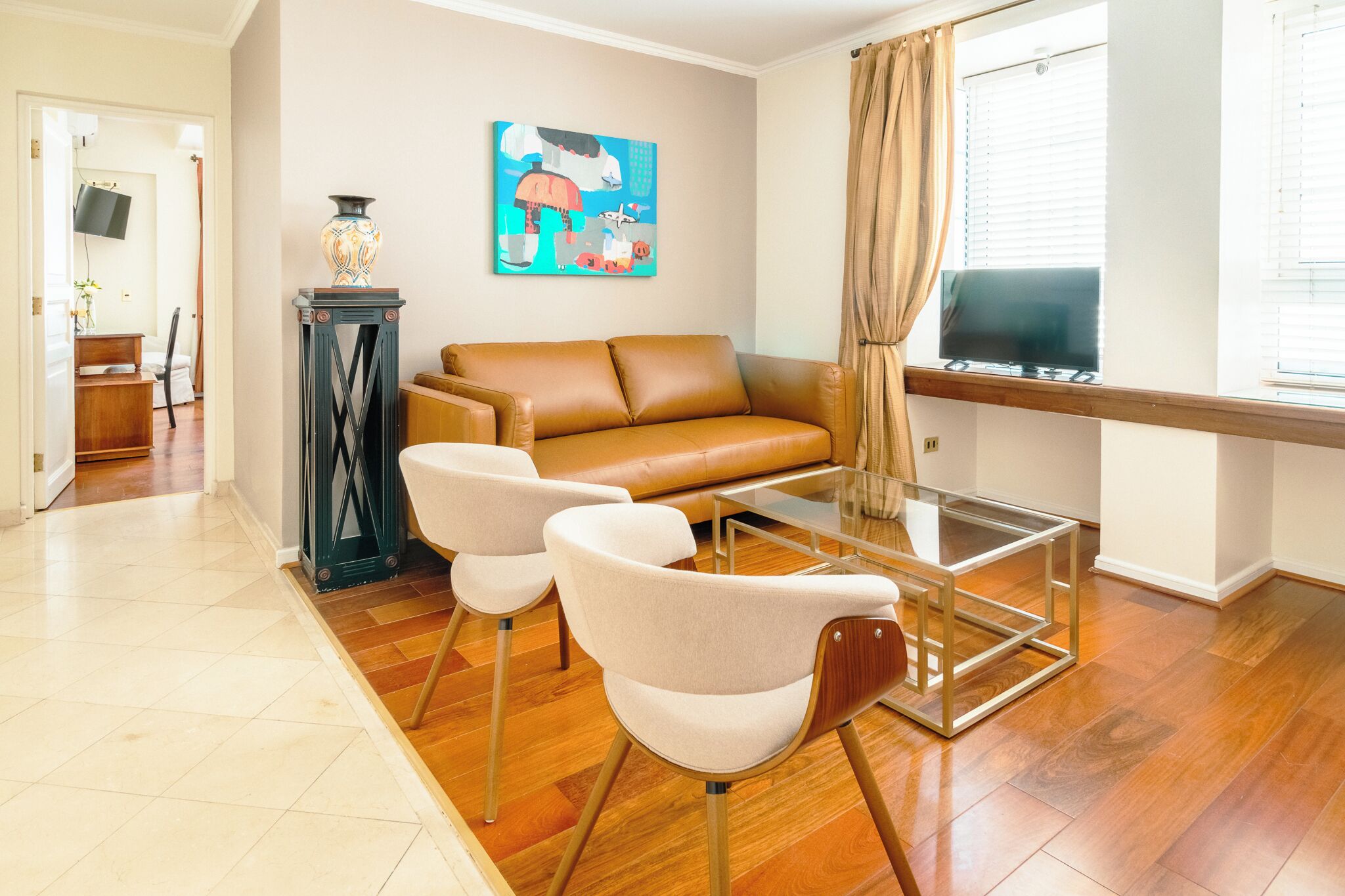 Una sala de estar con un sofá de cuero marrón, dos sillones blancos, una mesa de centro de vidrio, un jarrón decorativo y un cuadro de colores en la pared.