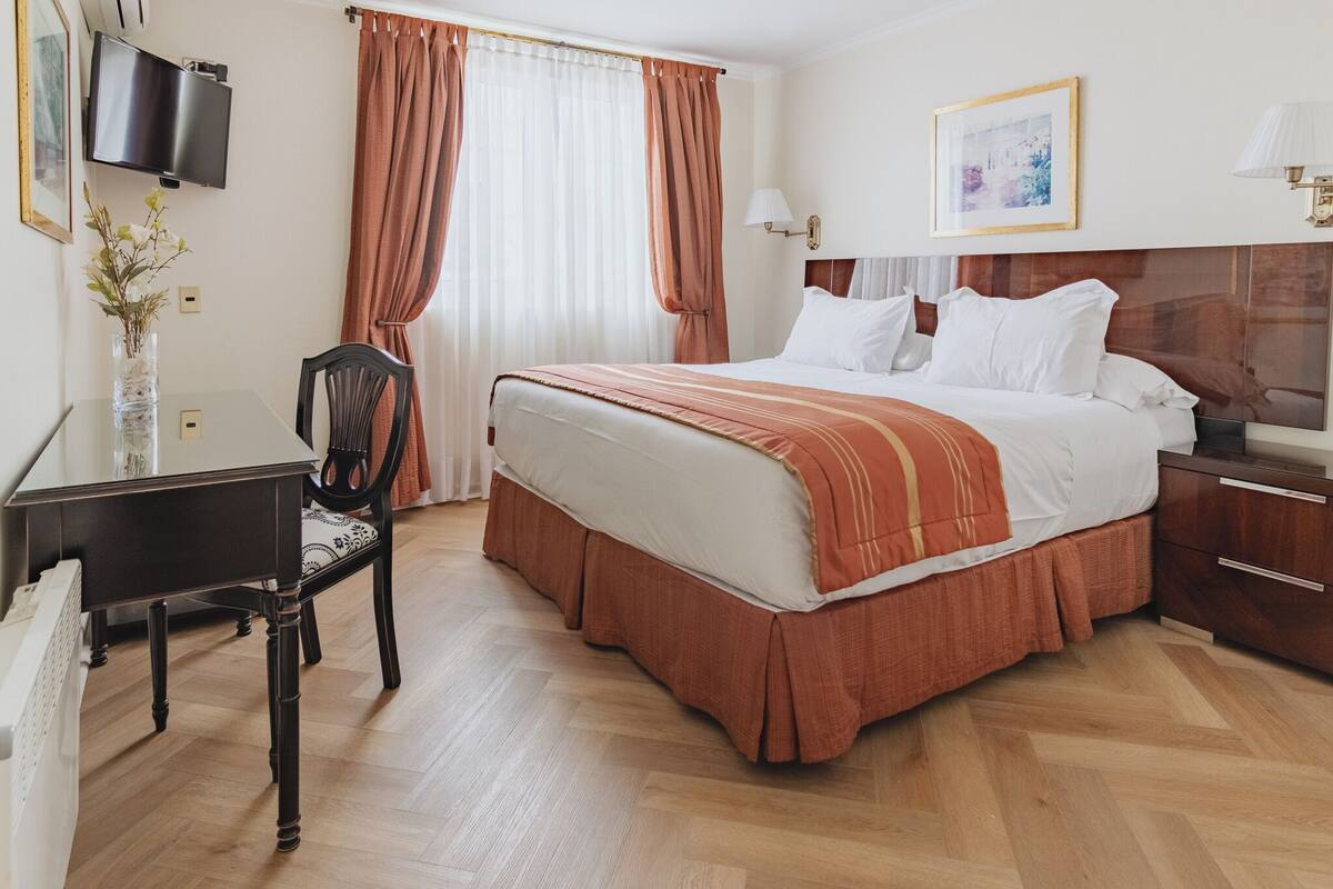 Ein Hotelzimmer mit einem großen Bett, einem Schreibtisch mit Stuhl, einem an der Wand montierten Fernseher und einem Fenster mit Vorhängen.