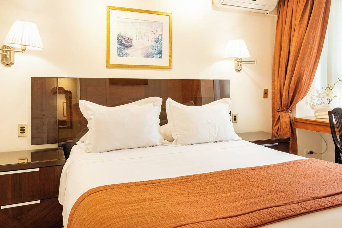 Ein Hotelzimmer mit einem großen Bett, zwei Nachttischen, einem Schminktisch, einem Spiegel, einem gerahmten Bild an der Wand und einem Fenster mit Vorhängen.