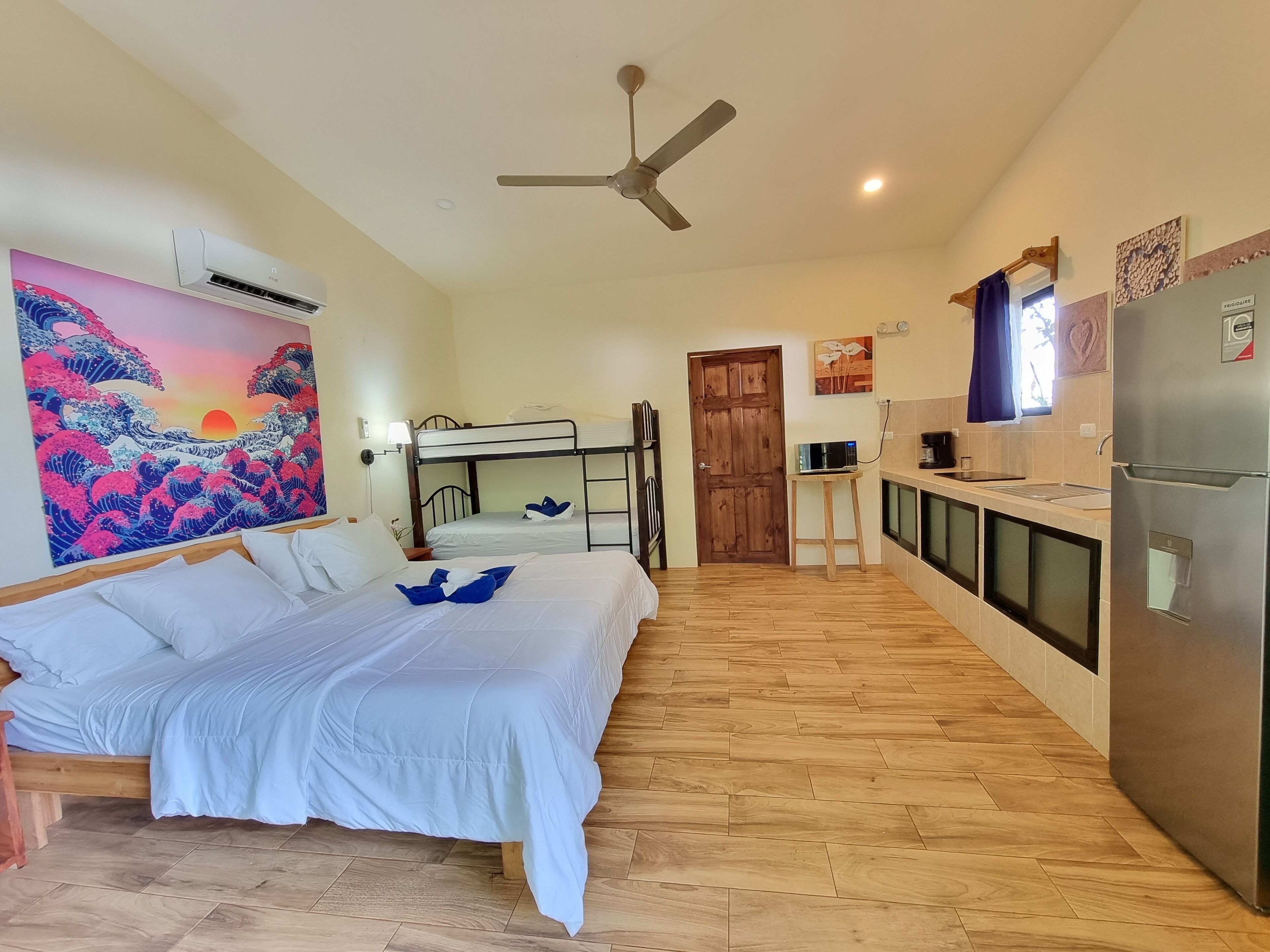 A room with bunk beds, a ceiling fan, an air conditioning unit, a kitchen with a refrigerator, and a wooden floor.