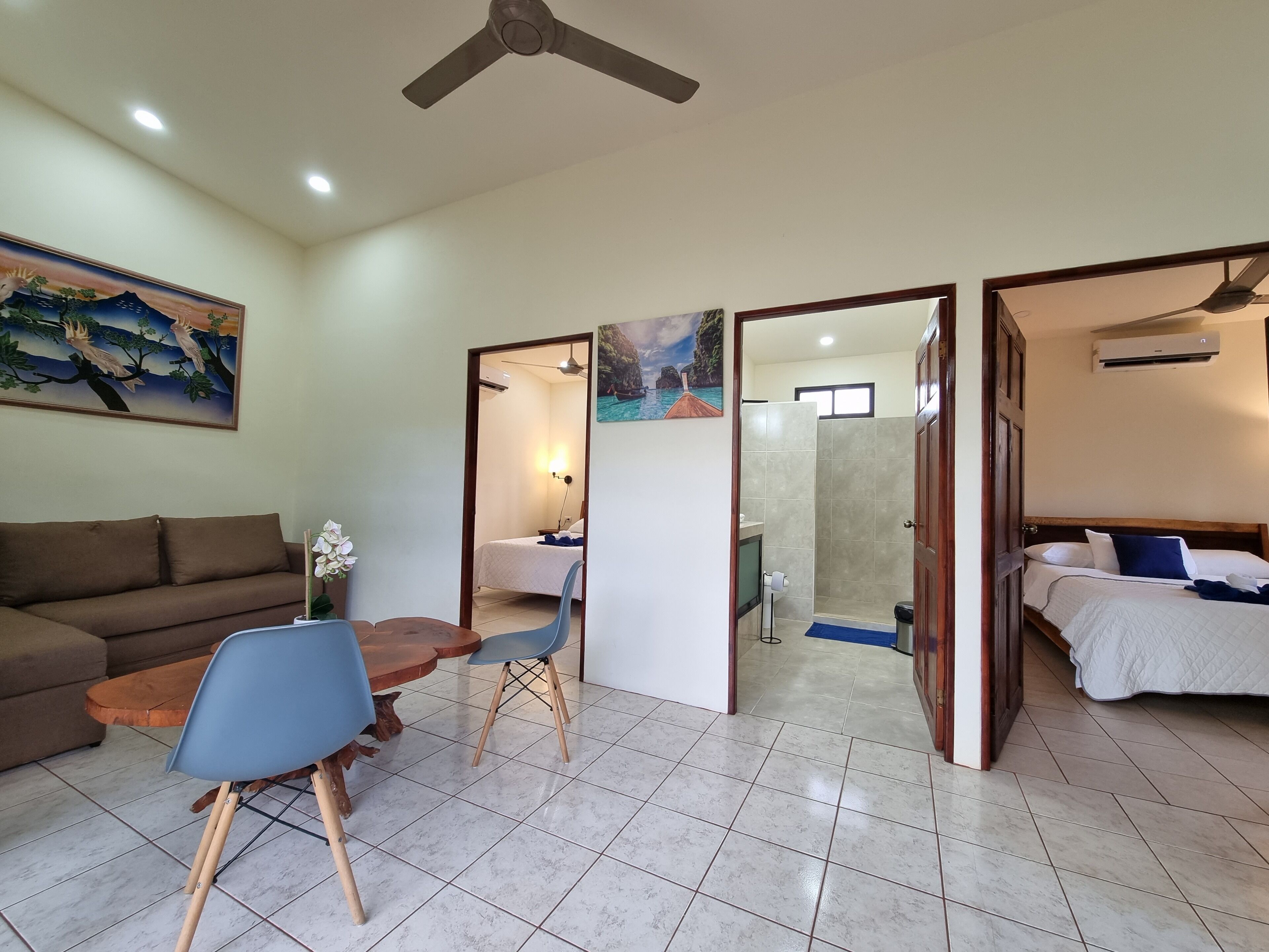 A hotel room with a bed, a sofa, a dining table with chairs, a ceiling fan, and a painting on the wall.