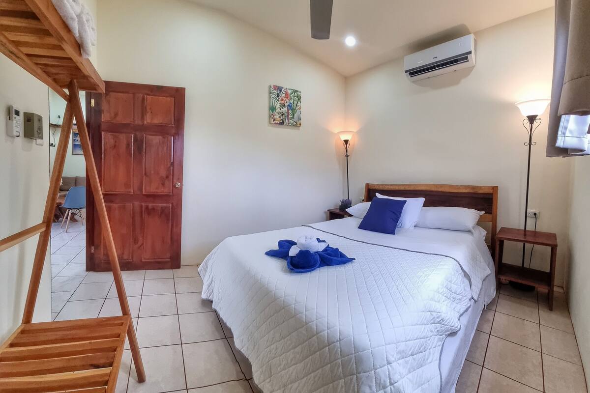 A hotel room with a bed, a wooden chair, a ceiling fan, an air conditioning unit, and a door.