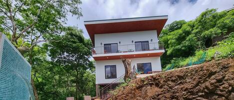 A modern two-story building with a swimming pool, surrounded by lush greenery and a rocky hillside.