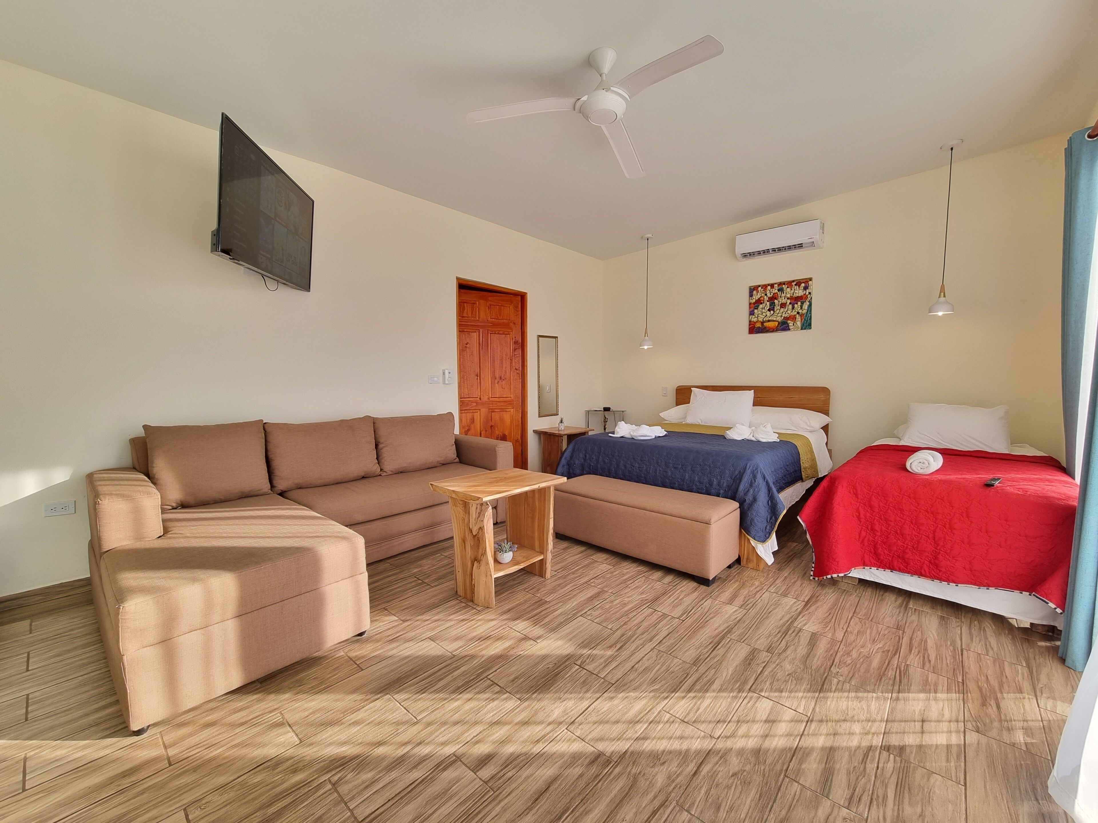 Een kamer met een bank, een bed met een rode deken, een houten tafel, een plafondventilator en een airconditioning.