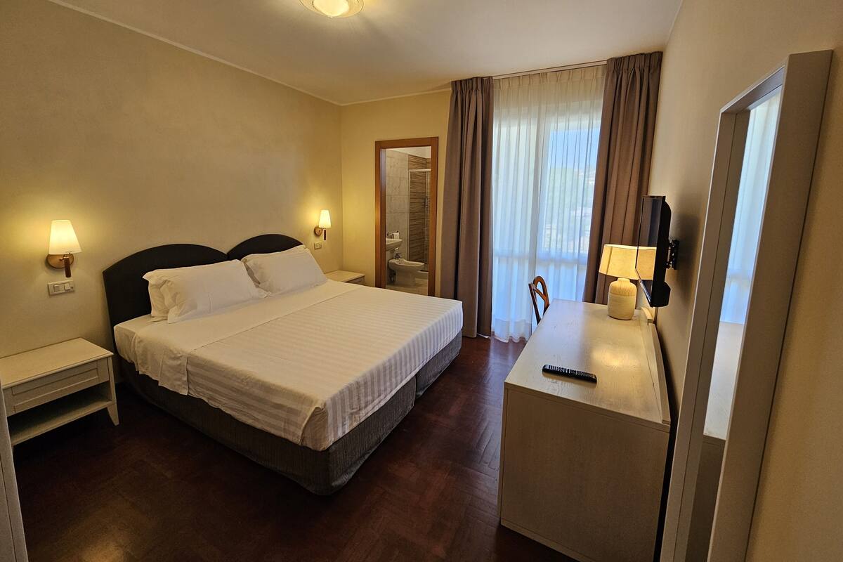 Een hotelkamer met een bed, nachtlampjes, een bureau met lamp, een televisie en een raam met gordijnen.