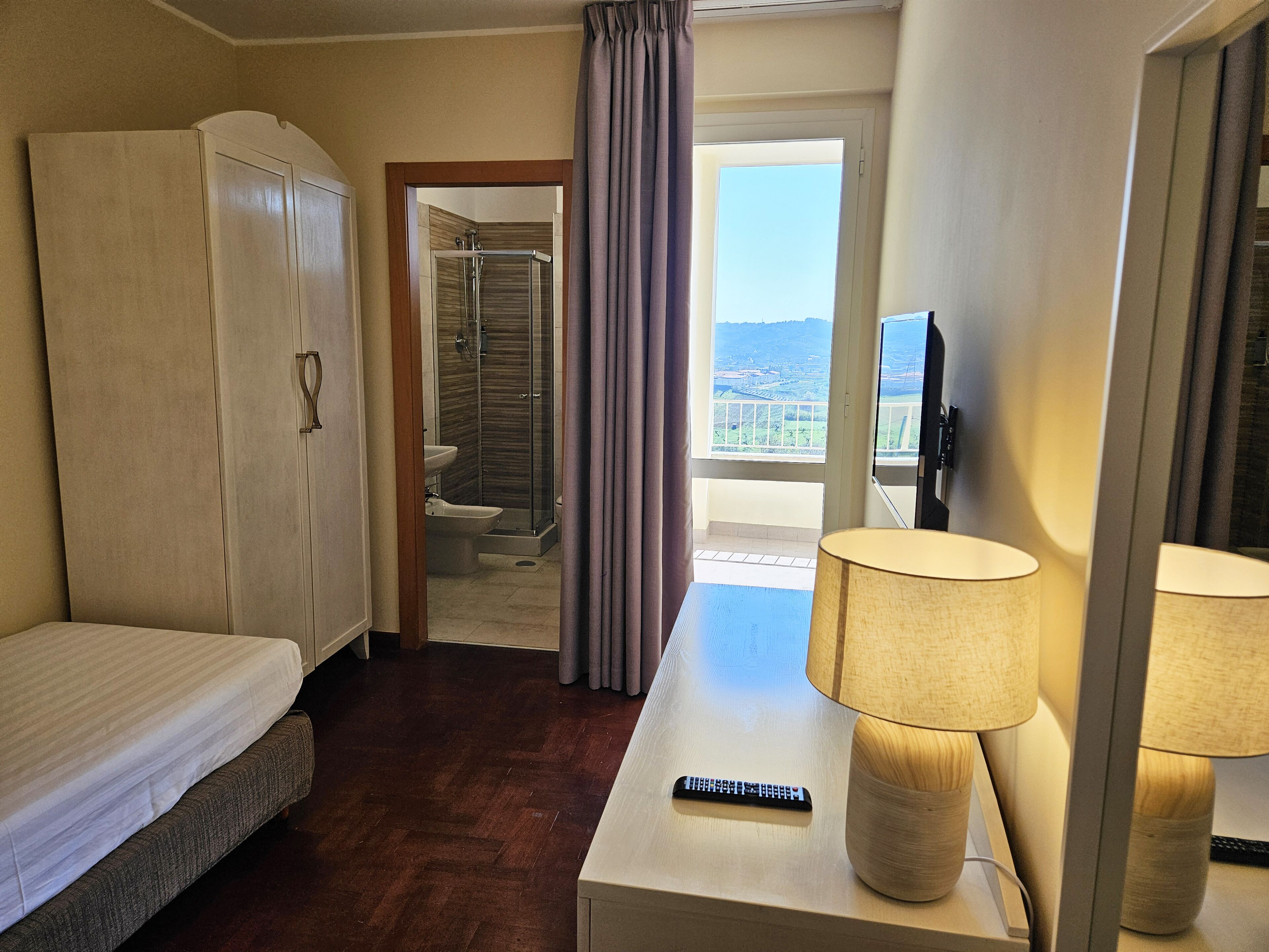 A hotel room with a bed, a desk with a lamp, a TV, a wardrobe, and a bathroom with a shower and toilet.