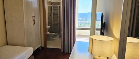 A hotel room with a bed, a desk with a lamp, a TV, a wardrobe, and a bathroom with a shower and toilet.