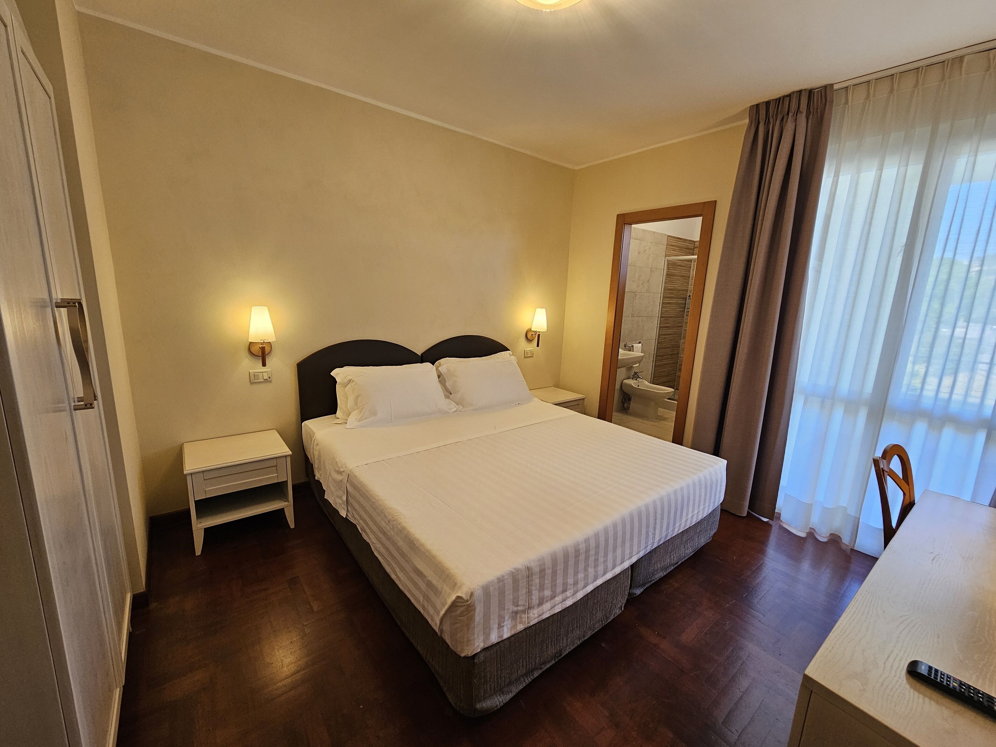 Habitación de hotel con una cama grande, lámparas de noche, una mesita, un espejo y vista al baño por una puerta.