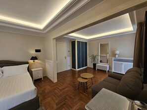 Een hotelkamer met een bed, een zithoek met een bank en een kleine tafel, een spiegel, een radiator en een deur naar een andere kamer.