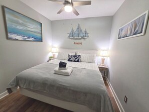 Quarto com cama, ventoinha no teto, mesa de cabeceira, luminária e um quadro de um veleiro na parede.