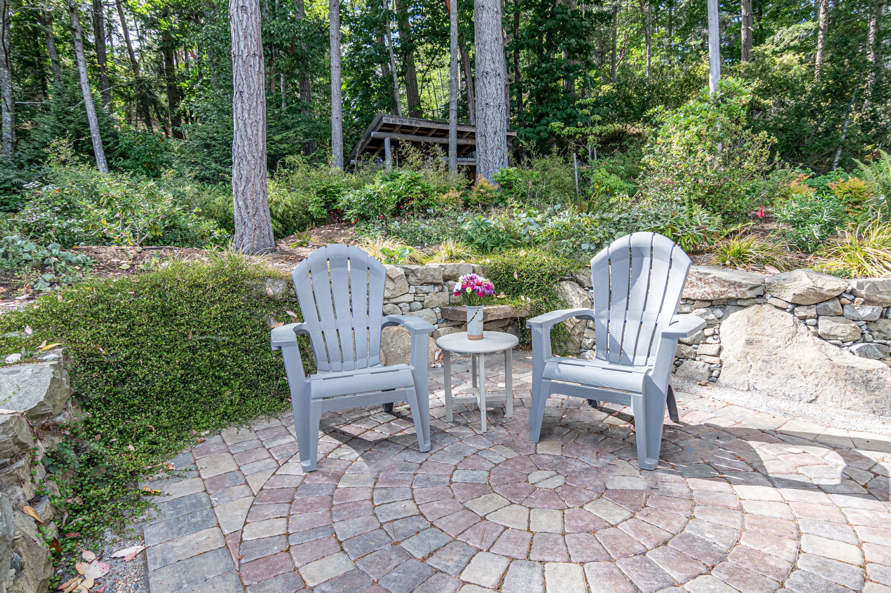 Two Adirondack chairs on a stone patio surrounded by lush greenery and a wooden pergola in the background.