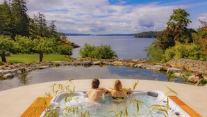 A hot tub with two people, surrounded by a stone wall and a wooden deck, overlooking a lake with trees and a distant shoreline. - Lightwater Cove (Salt Spring Island)