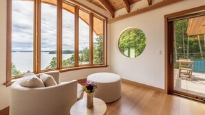 A room with a large window overlooking a lake, a round table with a vase of flowers, and a wooden ceiling. - Lightwater Cove (Salt Spring Island)