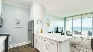 A modern kitchen with white cabinetry, a large island, and a view of the ocean through large windows. - 1205 Ocean Breeze Beachfront Retreat (Galveston)