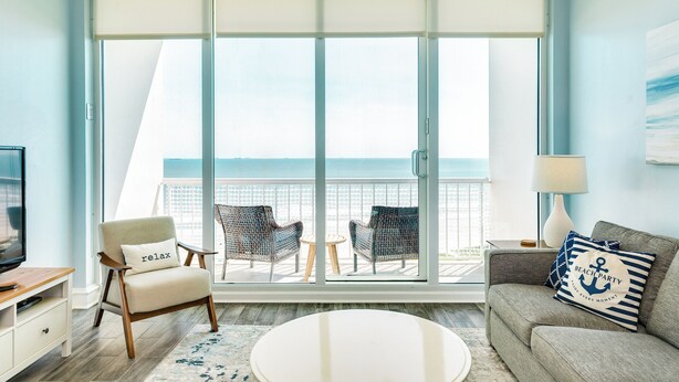 A room with a balcony, a sofa, a chair, a small table, and a TV. - 1205 Ocean Breeze Beachfront Retreat (Galveston)