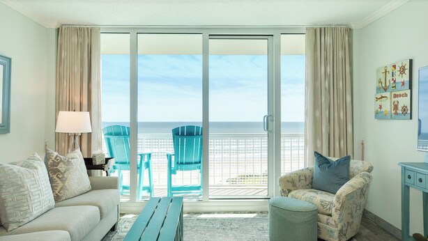 A room with a sofa, a chair, a small table, and a view of the beach through large glass doors.