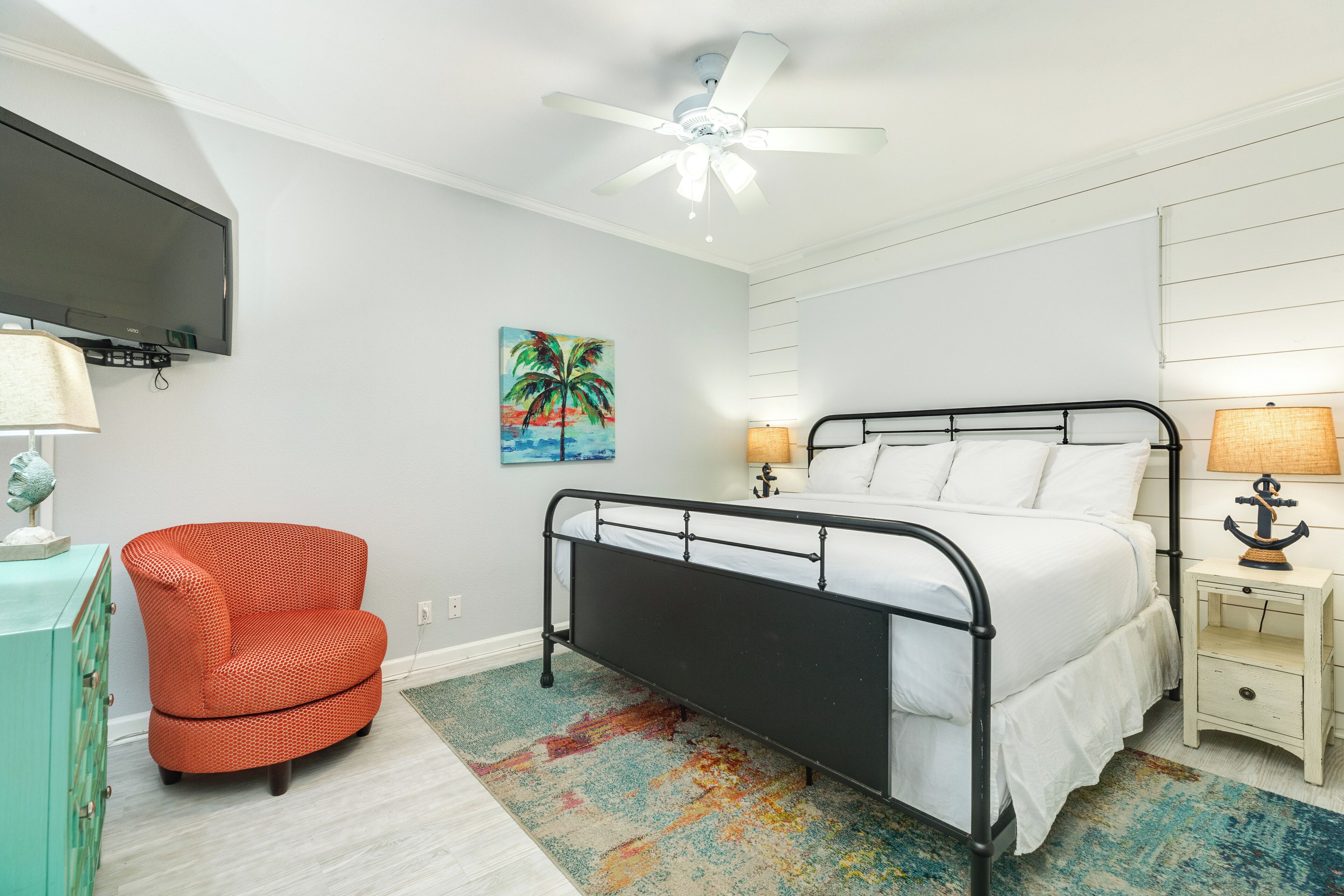 A bedroom with a bed, a chair, a TV mounted on the wall, and a ceiling fan.