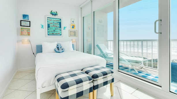 A bedroom with a bed, a bench, a glass door leading to a balcony with a view of the sea.