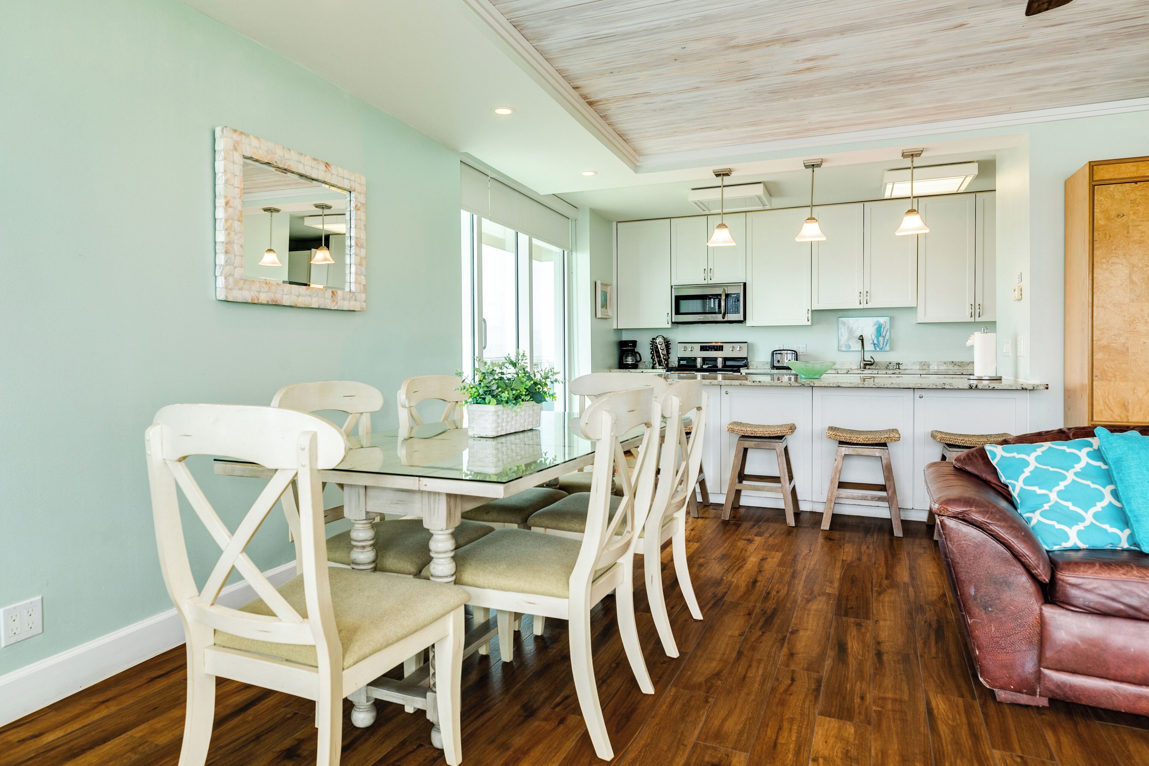 A dining area with a glass table and white chairs, a kitchen with white cabinets and a microwave, and a brown leather sofa with a patterned pillow.