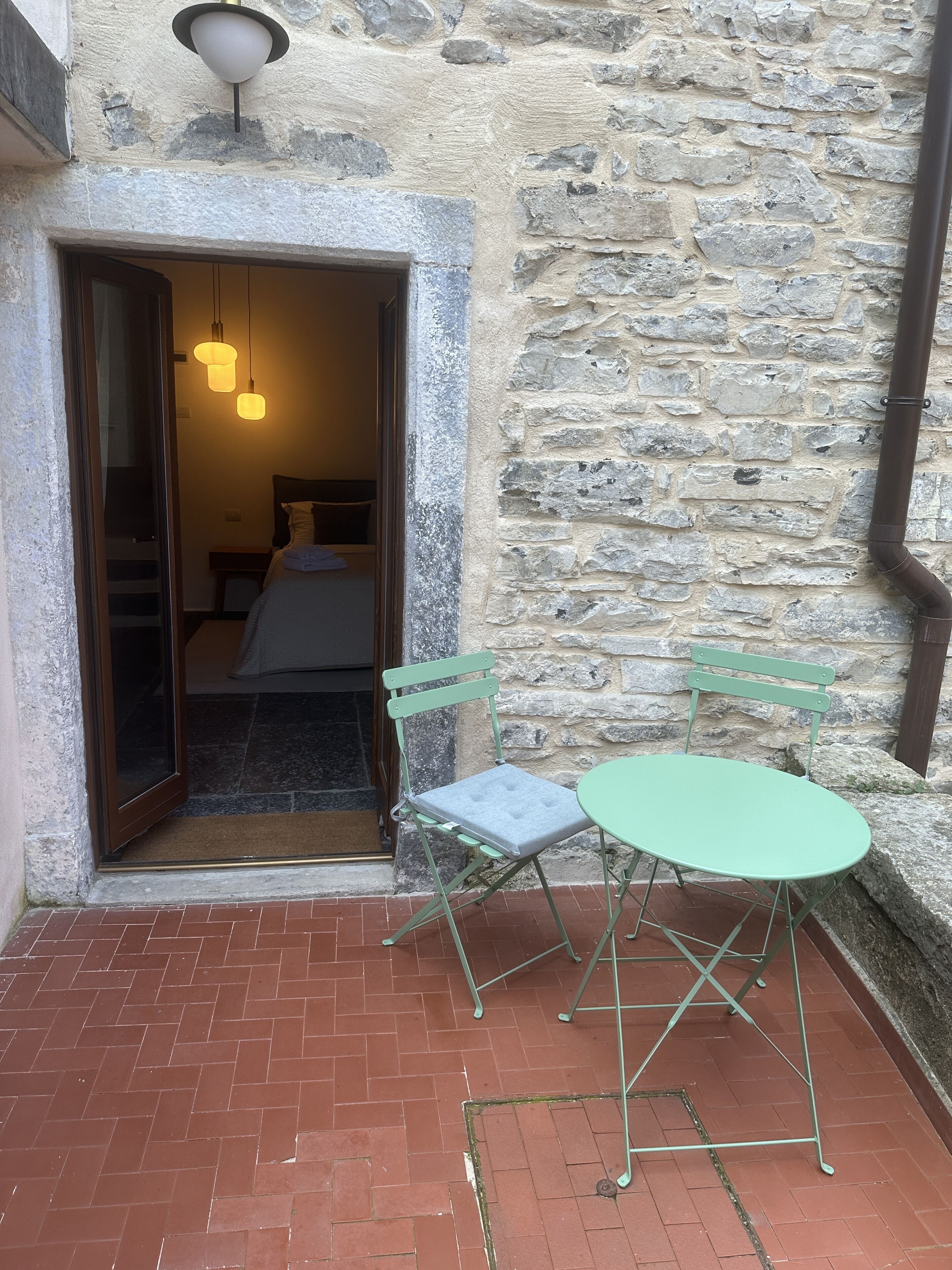 A stone-walled room with a bed, a green folding chair, and a small round table.