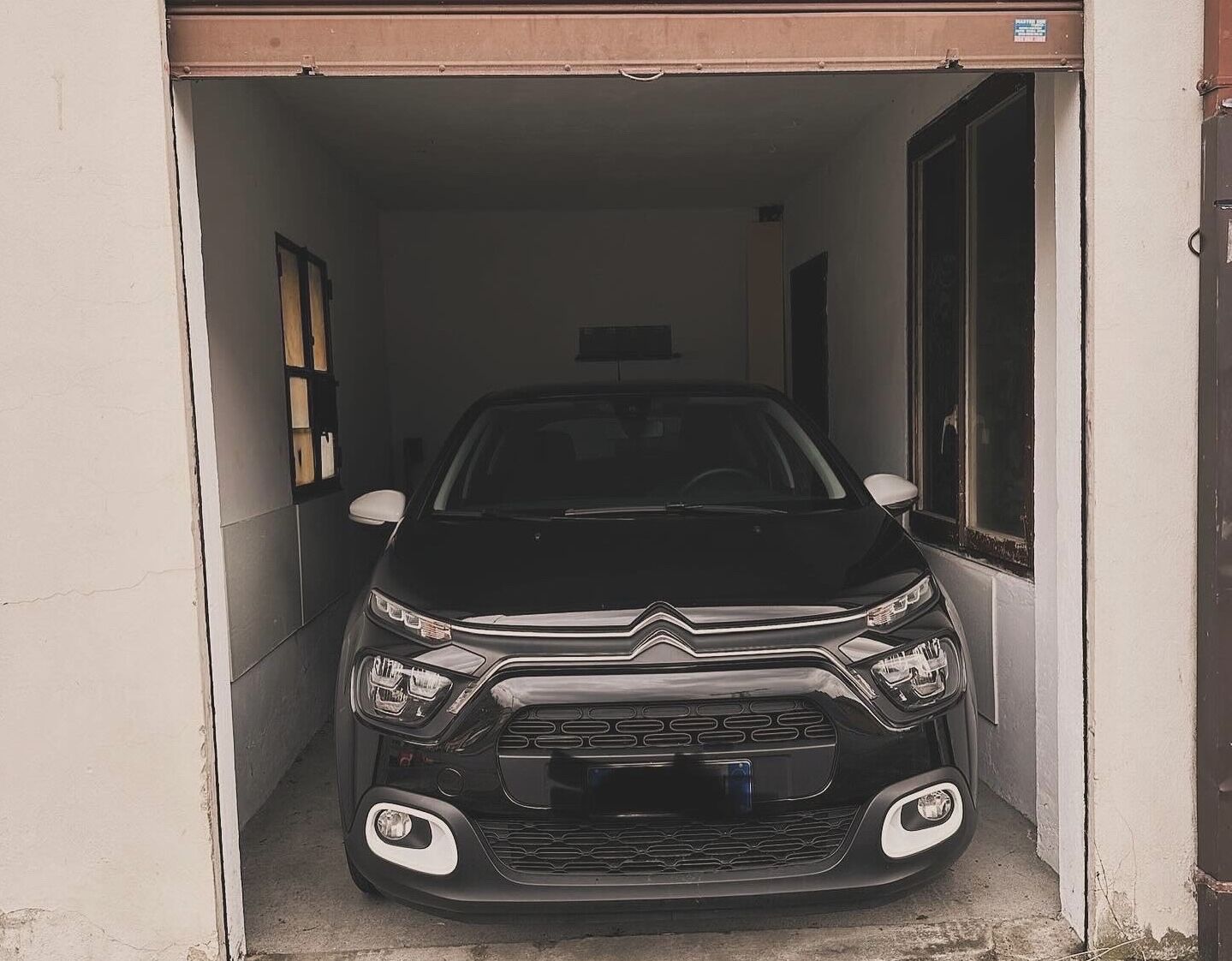 A black car parked inside a narrow, white-walled corridor with a door on the right and a window on the left.