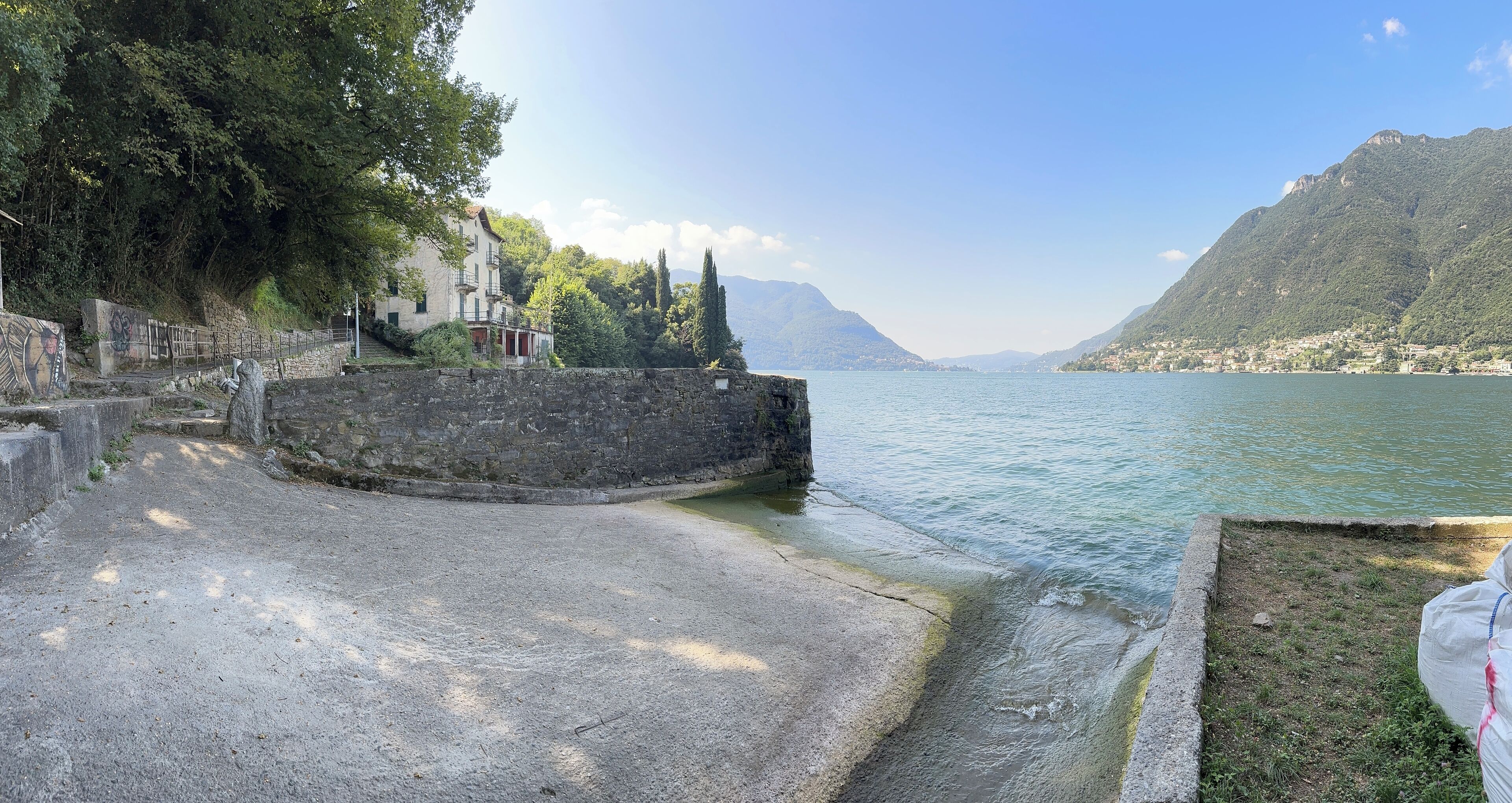 Ein Uferweg mit Steinmauer, Bäumen und Blick auf See und Berge.