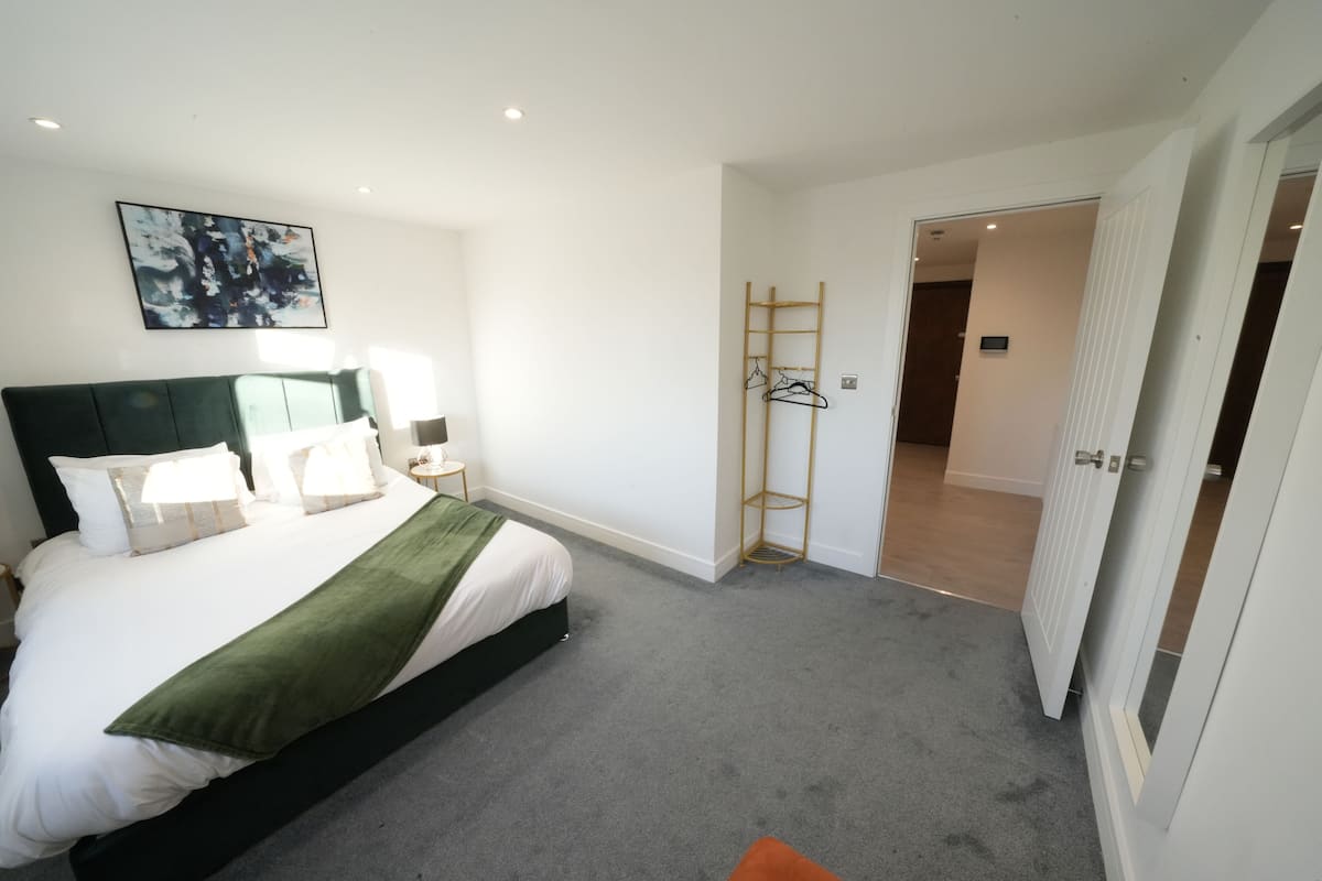 A bedroom with a bed, a bedside table, a lamp, a green throw blanket, a wooden ladder, and a door leading to another room.