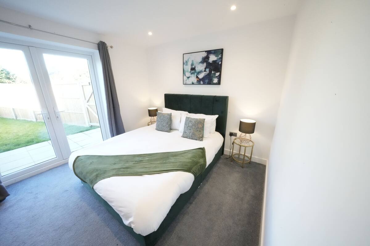 A bedroom with a large bed, a green throw, two bedside lamps, and a painting on the wall.