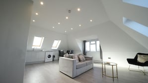 A spacious living area with a sofa, a round coffee table, and a black chair. There is a kitchen area with a washing machine and a dining table. The room has a sloped ceiling with skylights. - Heathrow Charge and Go (Ashford)