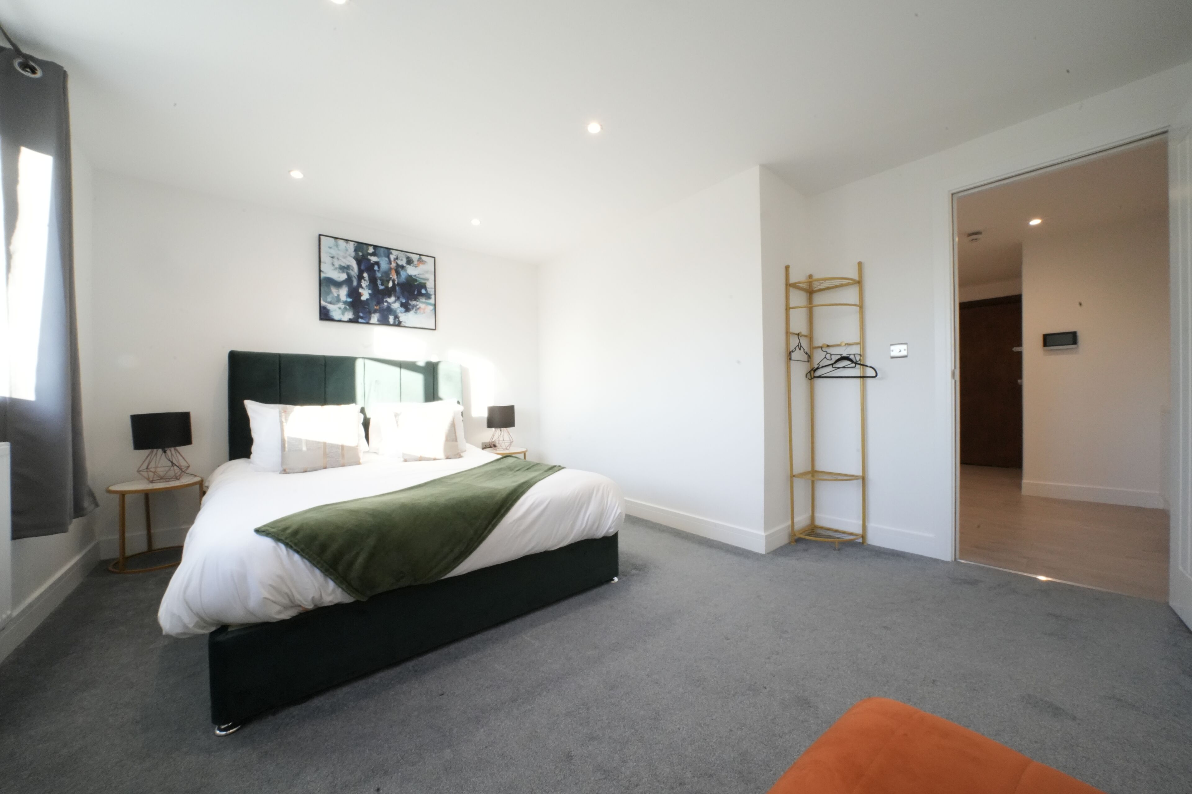 A modern hotel room with a large bed, a bedside table with a lamp, a green throw blanket, and a wooden ladder against the wall.