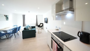 A modern kitchen with a black induction cooktop, a coffee maker, and a small dining area with blue chairs and a green sofa. - Heathrow Charge and Go (Ashford)