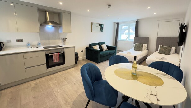 A modern hotel room with a kitchen, two beds, a dining area, and a large window. - Heathrow Charge and Go (Ashford)