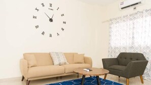 Una sala de estar con un sofá color beige, una mesa de centro redonda de madera, un sillón gris y un tapete azul estampado. Se observa un reloj de pared con números romanos.