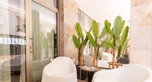 Un moderno vestíbulo de hotel con un puerta de vidrio que da a un balcón, una zona de estar con sillas blancas y una planta grande en maceta.
