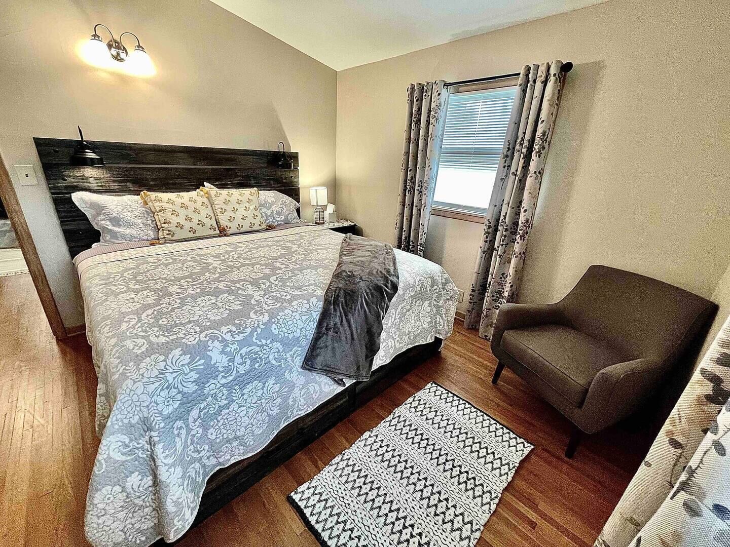 A bedroom with a large bed, a chair, a nightstand, and a window with curtains.
