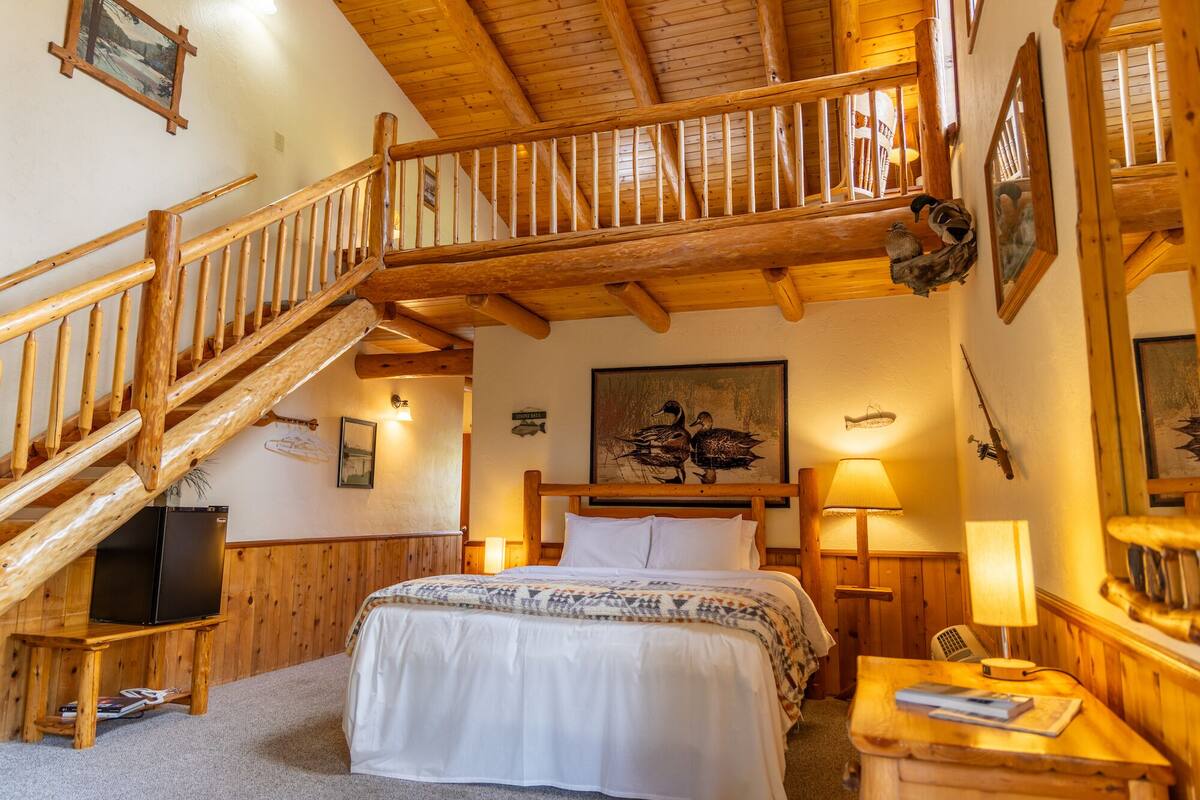 Een houten trap die leidt naar een slaapkamer op een entresol, met een bed, een bureau en een lamp.
