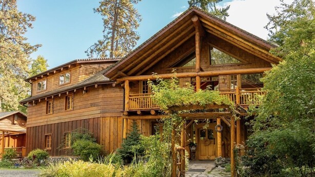 Une cabane en rondins dotée d’un balcon, d’un chemin de pierre et entourée de verdure.