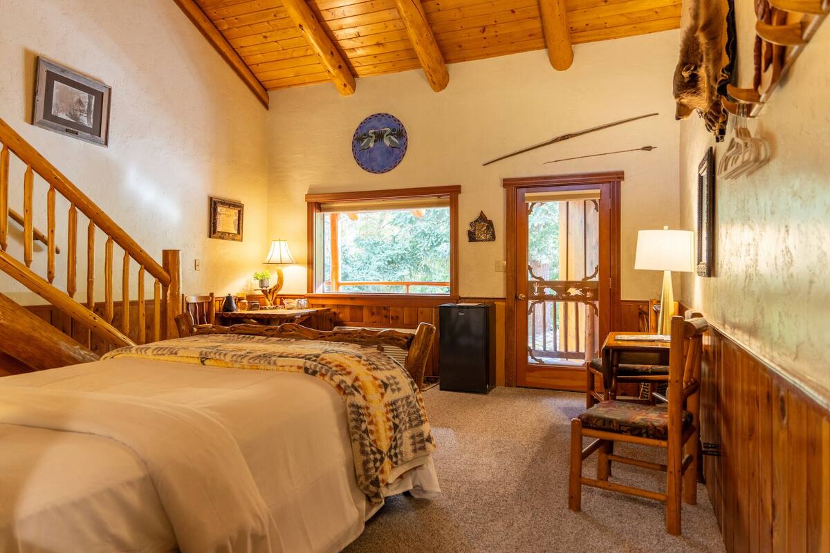 Een knus ingerichte kamer met een houten bed, een bureau, een stoel en een raam met uitzicht op bomen.