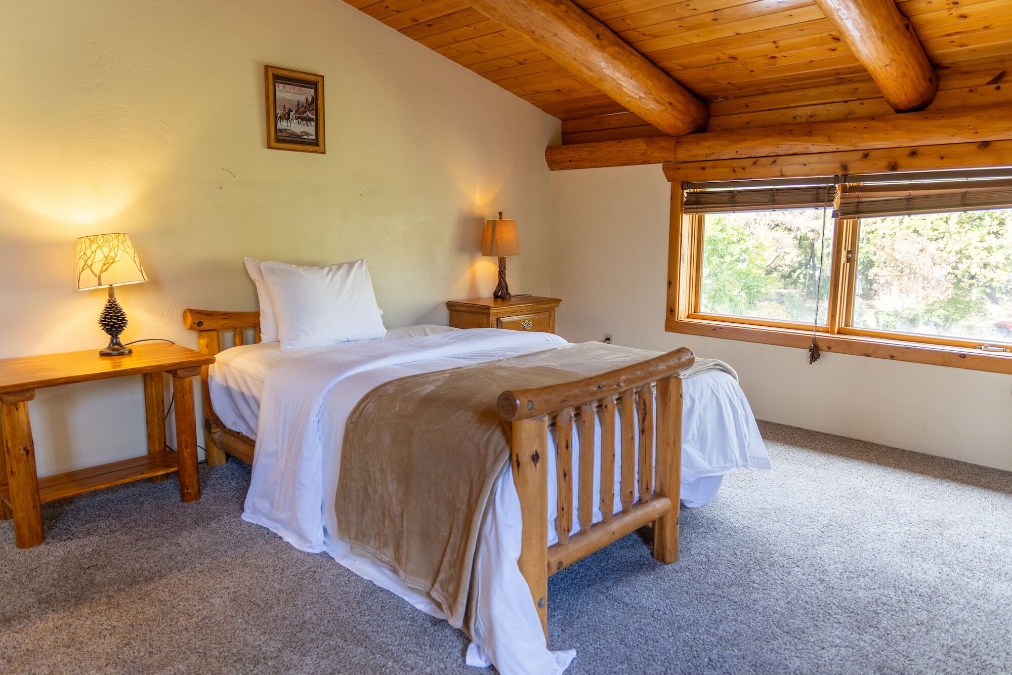 A cozy bedroom with a wooden bed, two bedside tables with lamps, a window with blinds, and a framed picture on the wall.