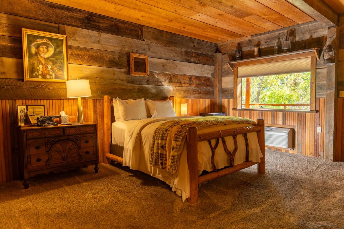 Een eenvoudige, houten slaapkamer met een bed, nachtkastje, lamp en raam.