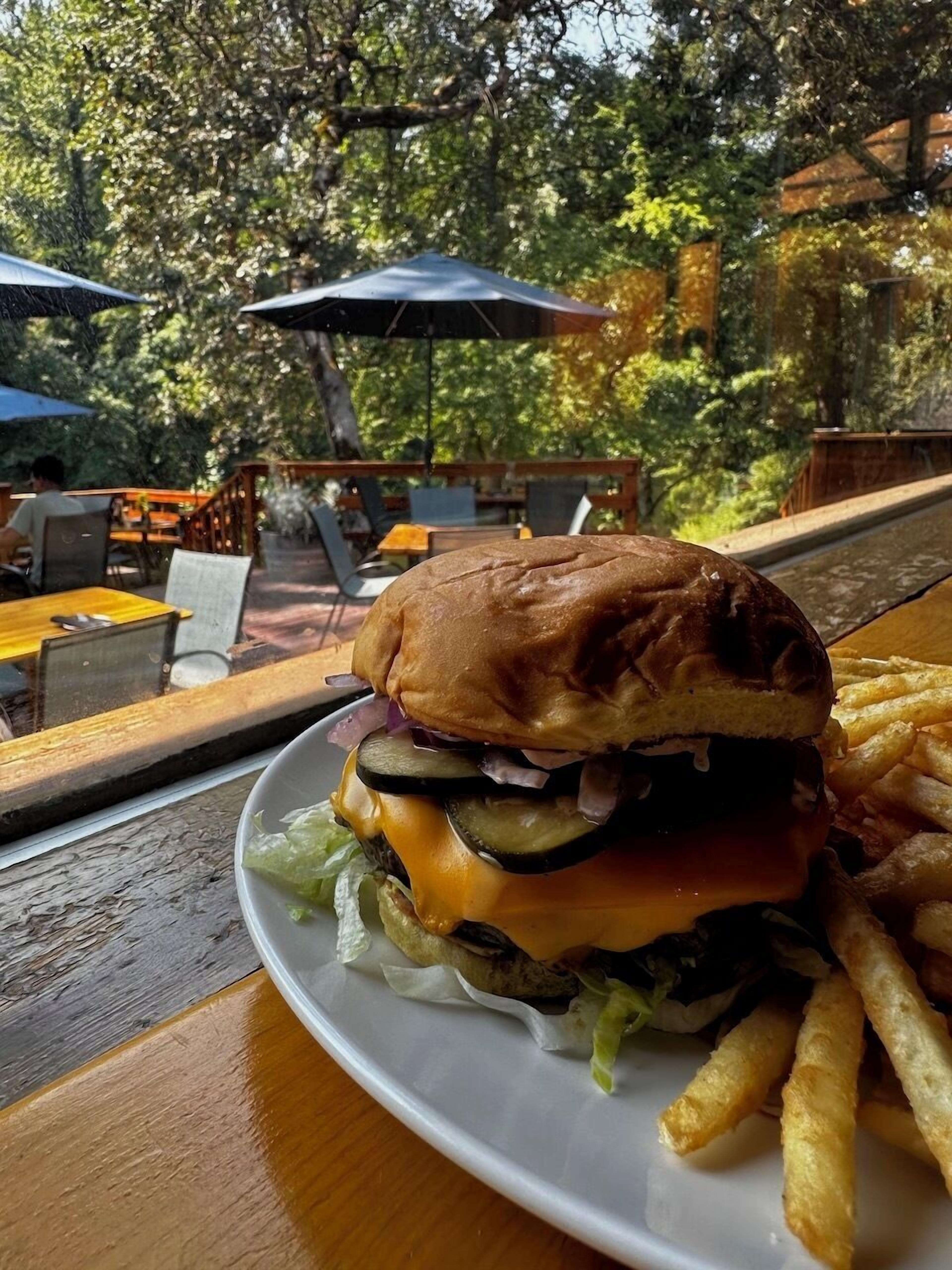 En hamburgare med pommes frites på ett träbord i en utomhusmiljö med stolar och parasoller.