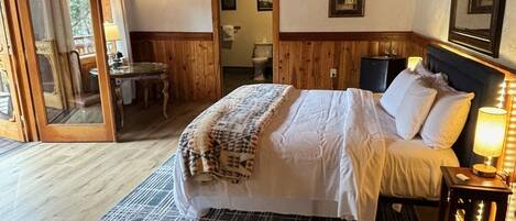 A wooden-paneled bedroom with a bed, a nightstand, a mirror, and a door leading to a bathroom.